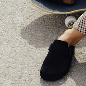 Person wearing black clogs standing on a skateboard with a checkered shirt sleeve.